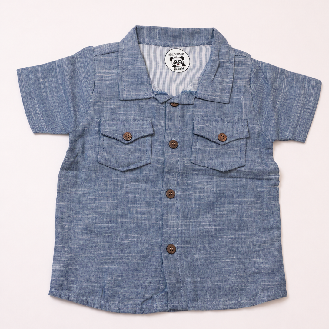 A short-sleeve baby shirt in a textured blue chambray fabric, featuring a camp collar, brown buttons, and two chest pockets,